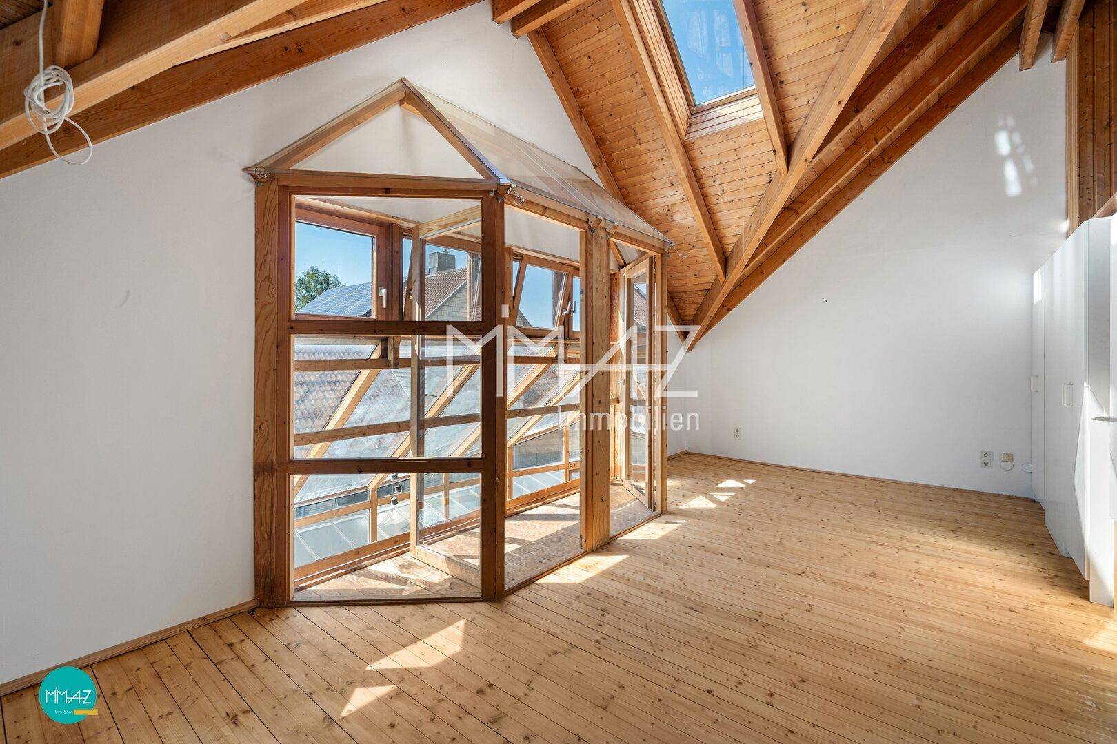 Geräumiges Dachzimmer mit Holzbalken, Dielenboden und einer verglasten Nische.