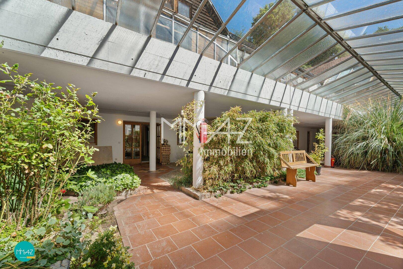 Überdachte Terrasse mit Fliesenboden, Sitzbank und üppiger Bepflanzung.