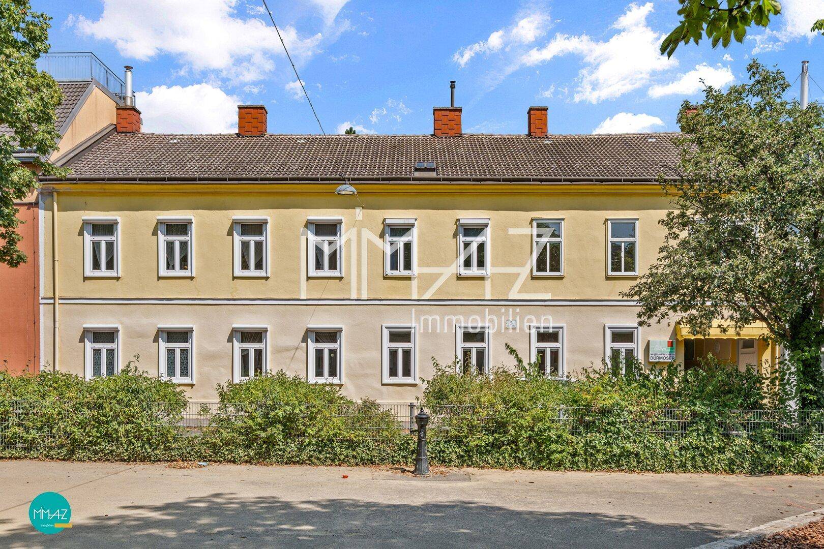 Vorderfassade des gelben Mehrfamilienhauses mit vielen Fenstern und grünen Büschen.