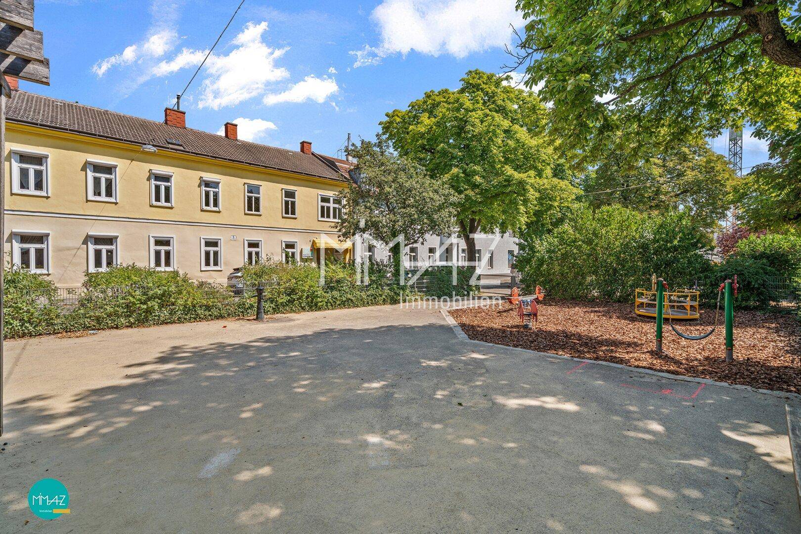 Blick auf eine Wohnstraße mit einem gelben Gebäude, Bäumen und einem kleinen Spielplatz im Vordergrund.