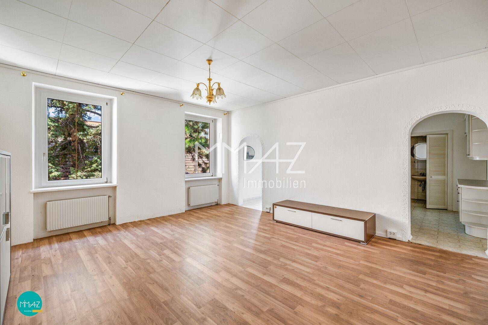 Helles Wohnzimmer mit Parkettboden, zwei Fenstern und einem Durchgang zu einem weiteren Bereich.