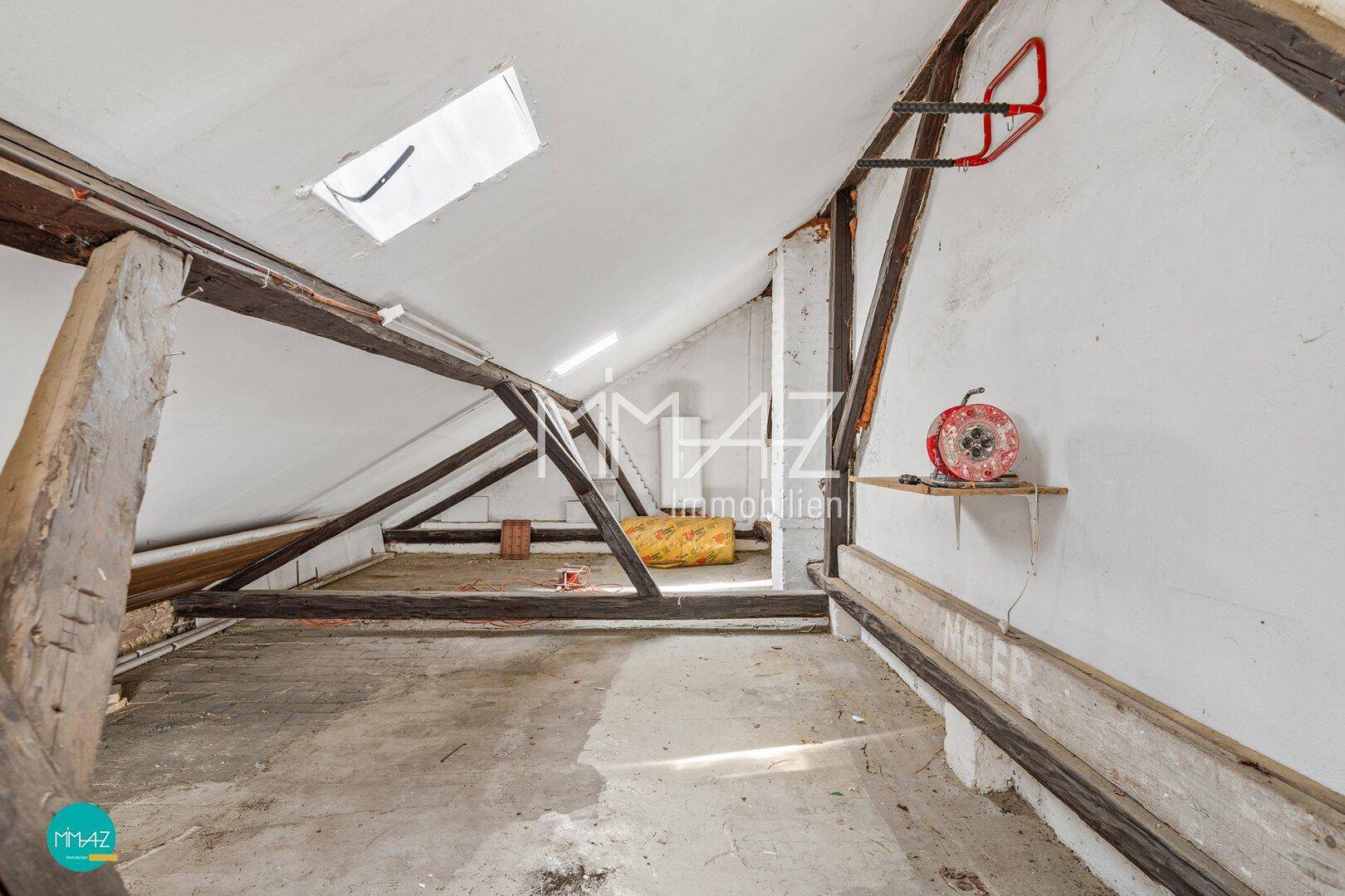 Unausgebauter Dachboden mit Holzbalken, Schrägen und einem Dachfenster für natürliches Licht.