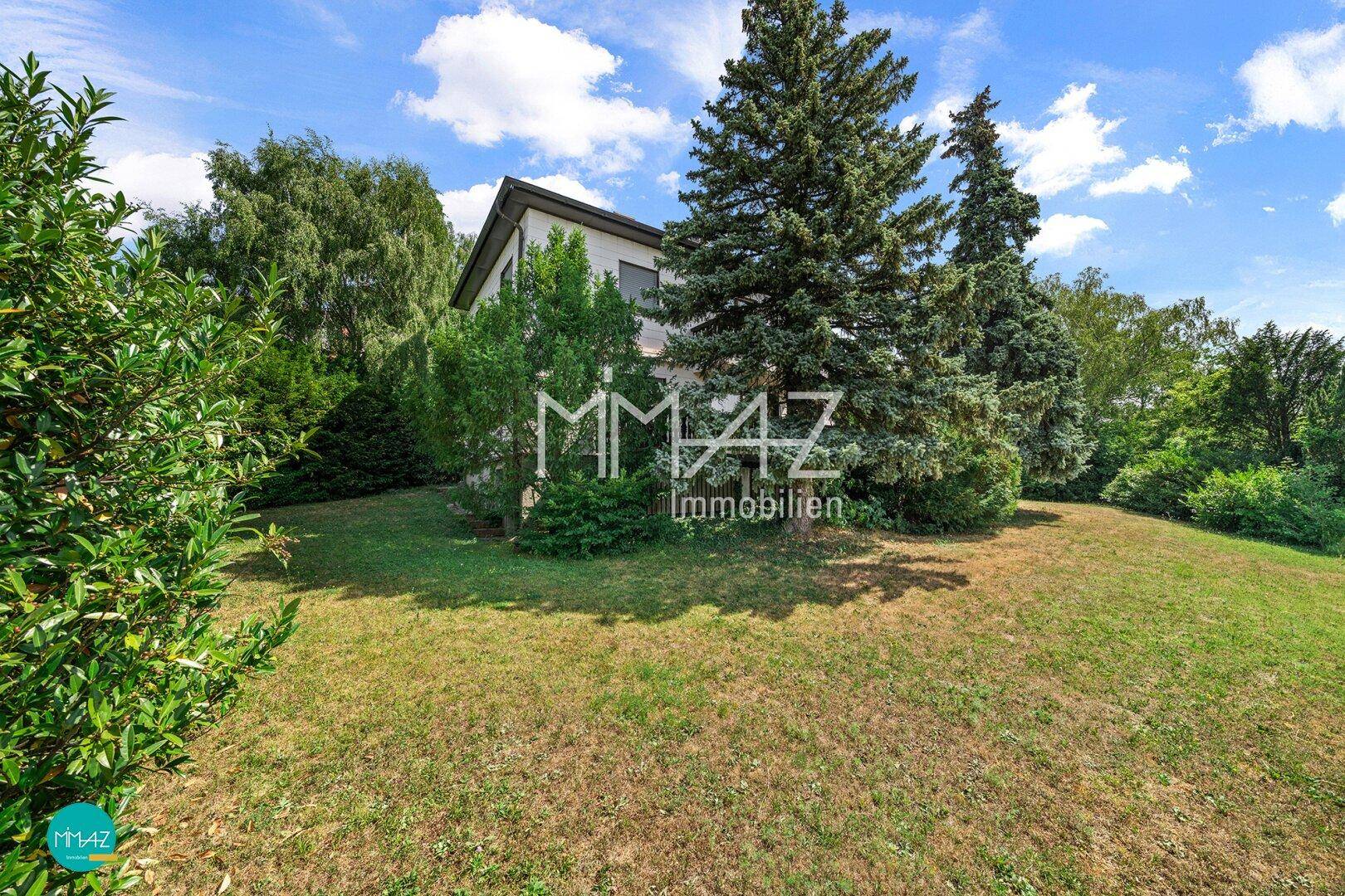 Das Haus umgeben von einem weitläufigen Garten mit hohen Nadelbäumen unter blauem Himmel.