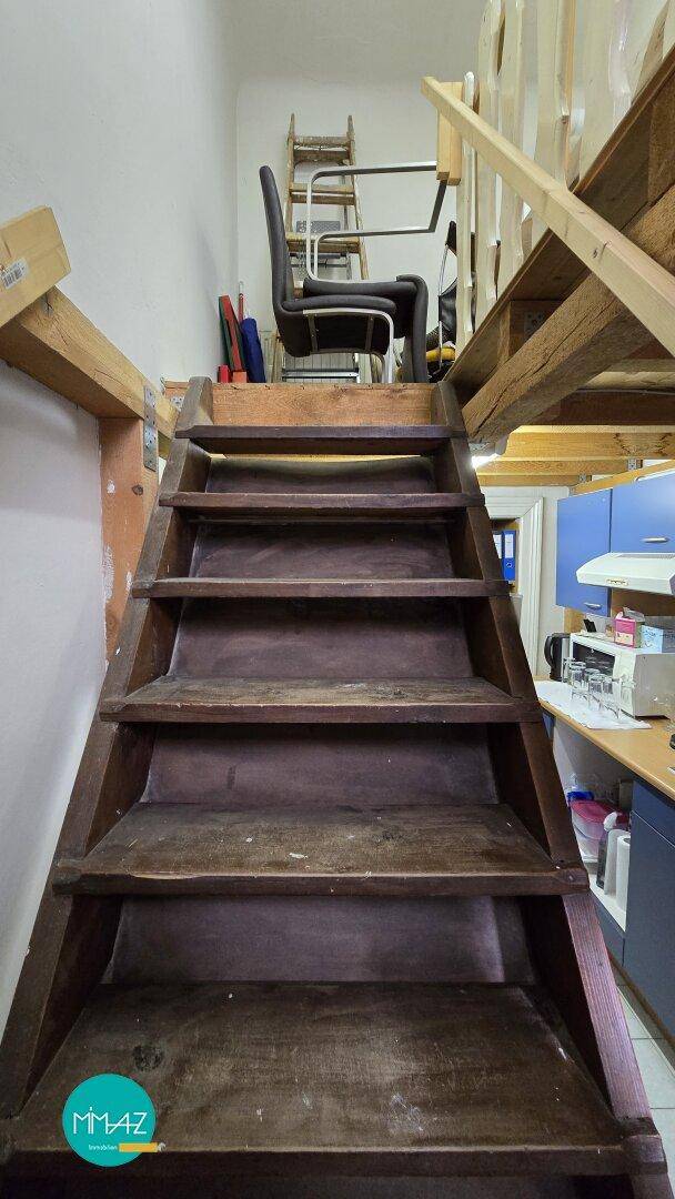 Steile Holztreppe führt zu einem Zwischengeschoss, mit sichtbaren Holzbalken und Lagerbereich.