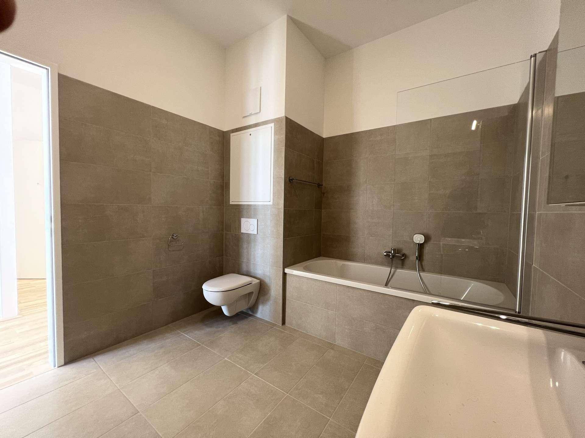 Helles Badezimmer mit Badewanne, wandhängendem WC und grauen Fliesen.