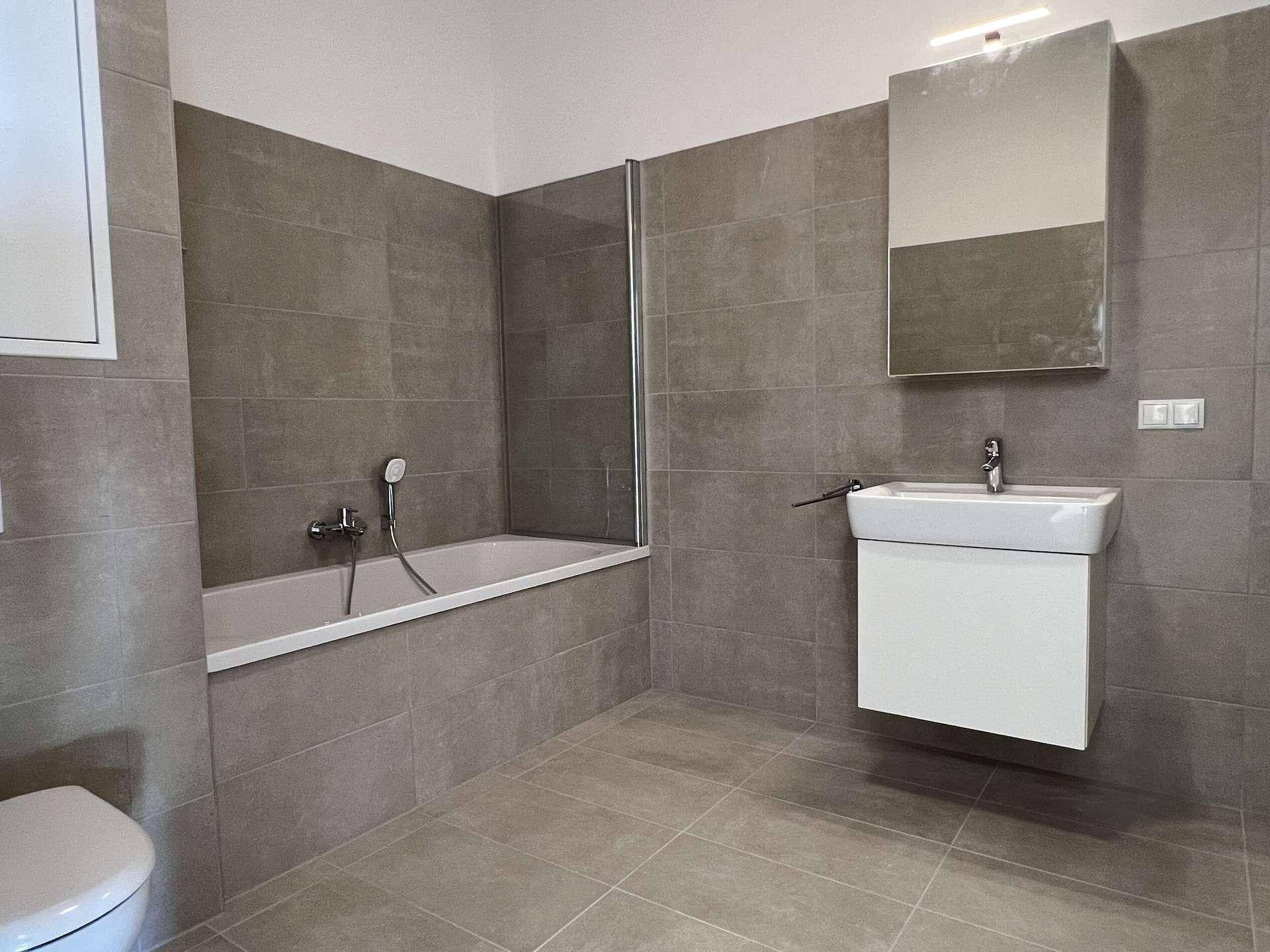 Modernes Badezimmer mit grauen Fliesen, Badewanne mit Duschwand und Waschbeckenunterschrank.
