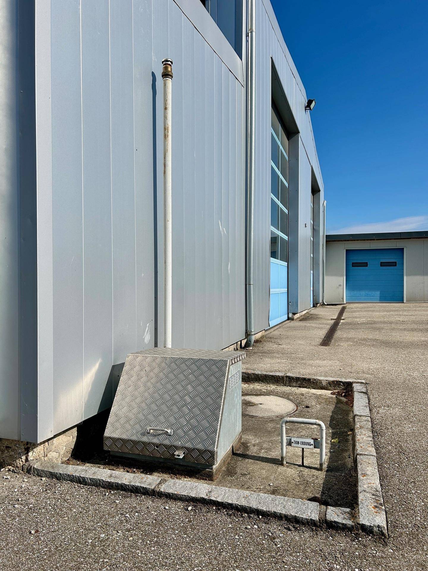 Detailansicht der Gebäudefassade mit Metallverkleidung, einem Entwässerungsrohr und einem Wartungsschacht im Außenbereich.