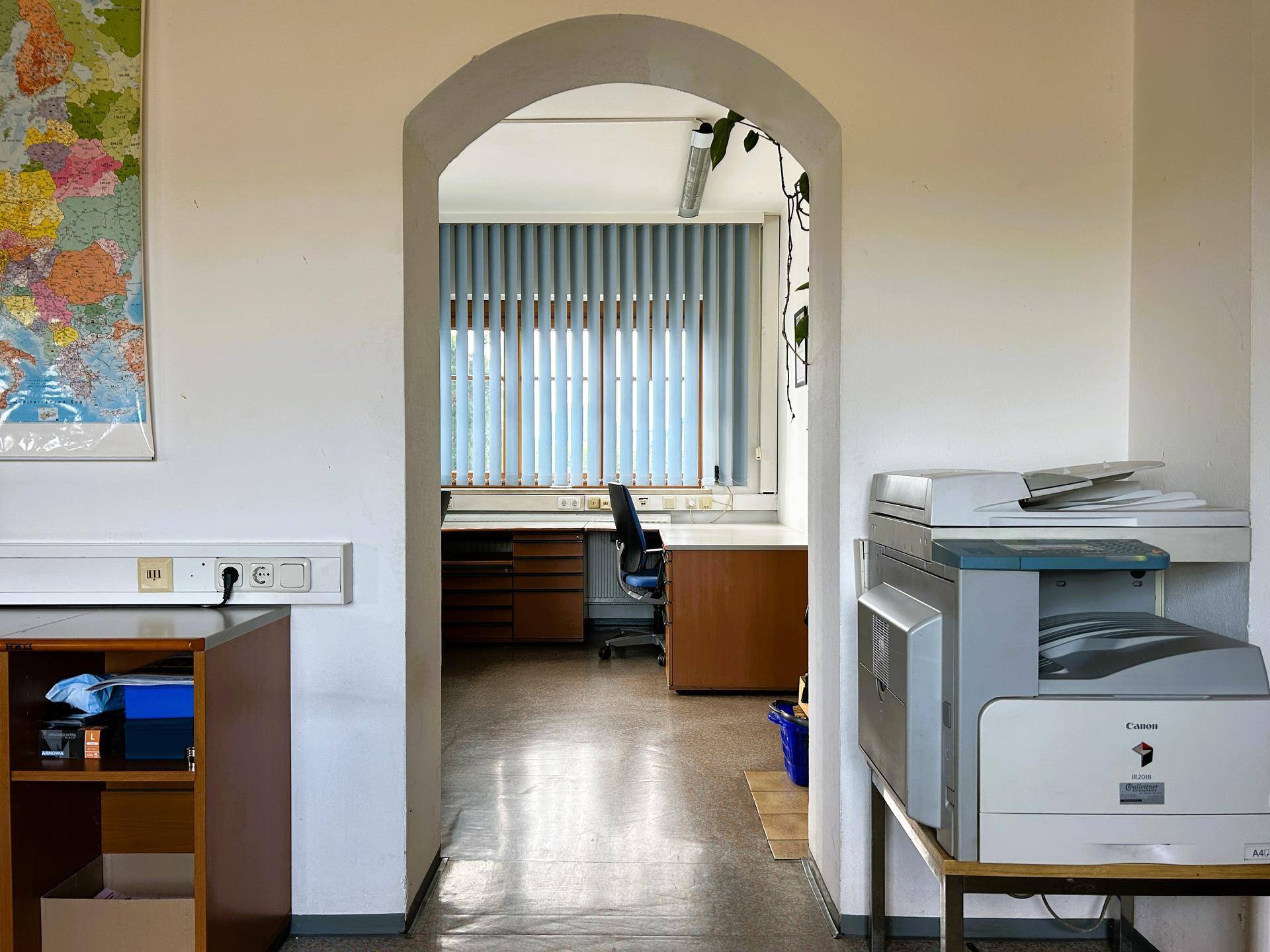 Blick durch einen Torbogen in einen weiteren Bürobereich mit Schreibtischen und Fenstern, die Tageslicht hereinlassen.