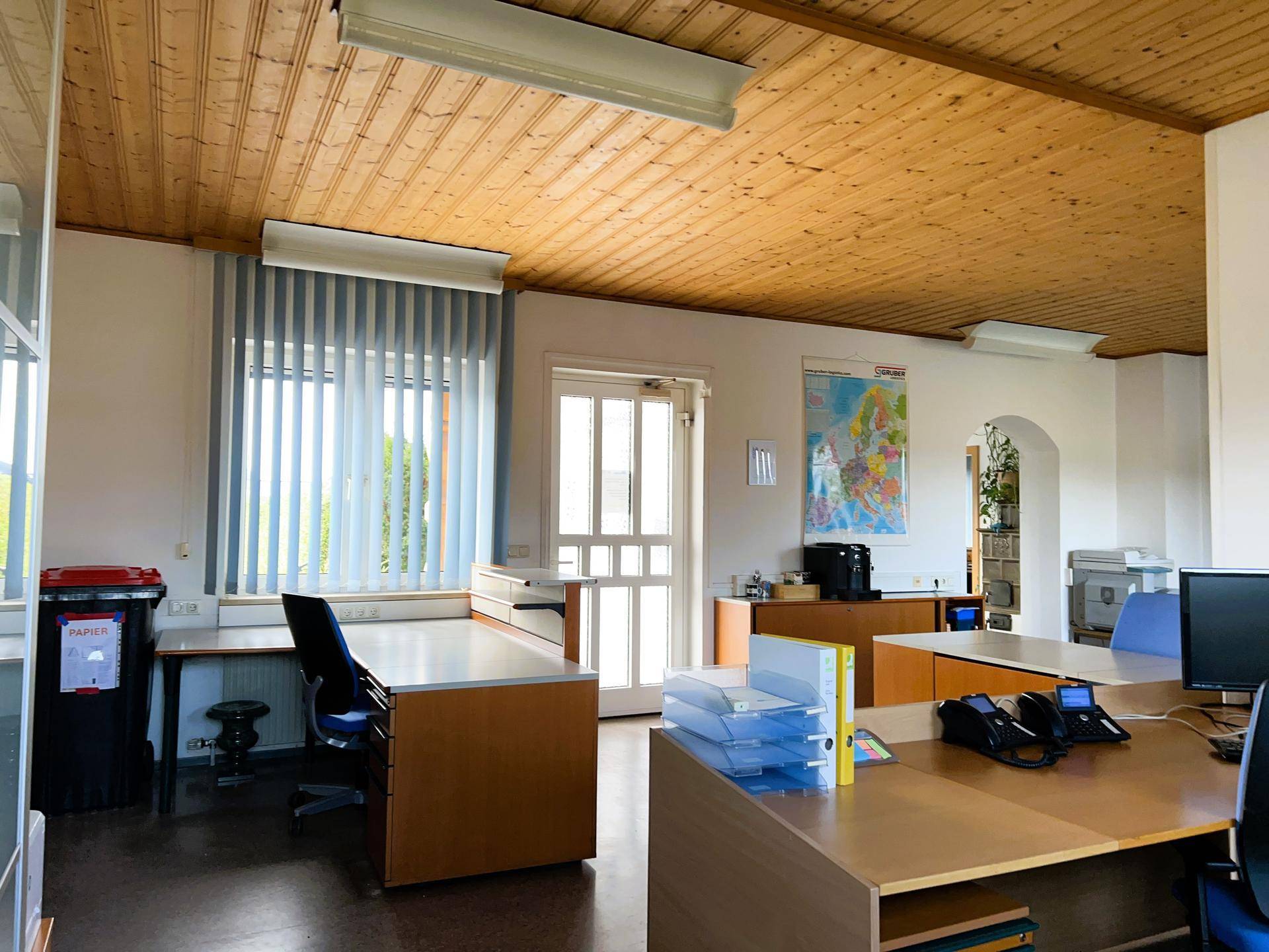 Helles Büro mit Holzdecke und vertikalen Jalousien an den Fenstern, ausgestattet mit mehreren Arbeitsplätzen.