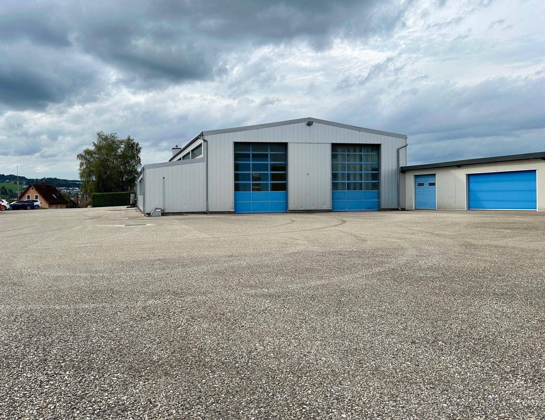 Gewerbegebäude mit blauen Toren und einem weitläufigen Vorplatz unter einem dramatischen, bewölkten Himmel.