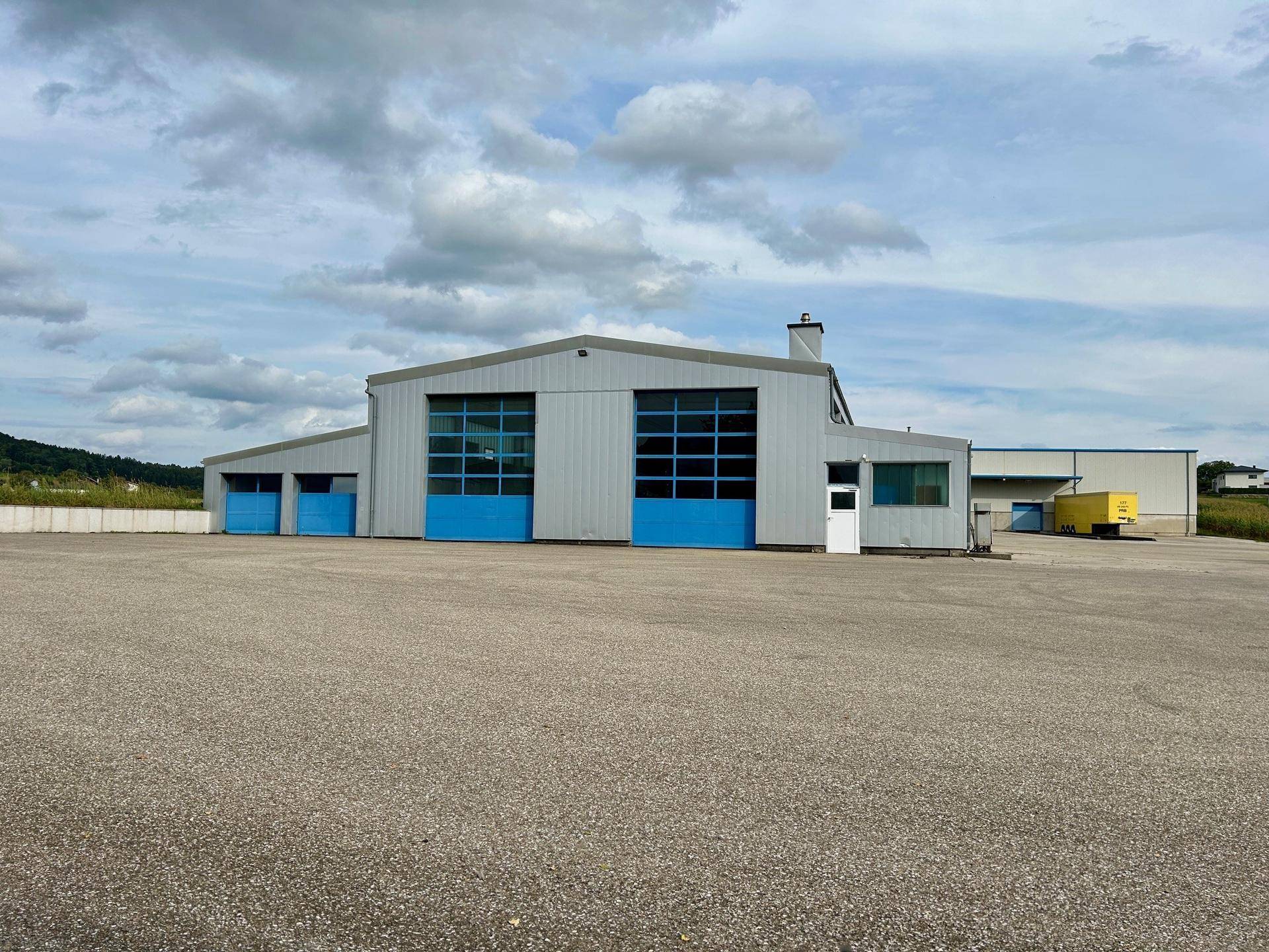 Gewerbegebäude mit blauen Toren und großem asphaltierten Vorplatz unter bewölktem Himmel, umgeben von grüner Landschaft.