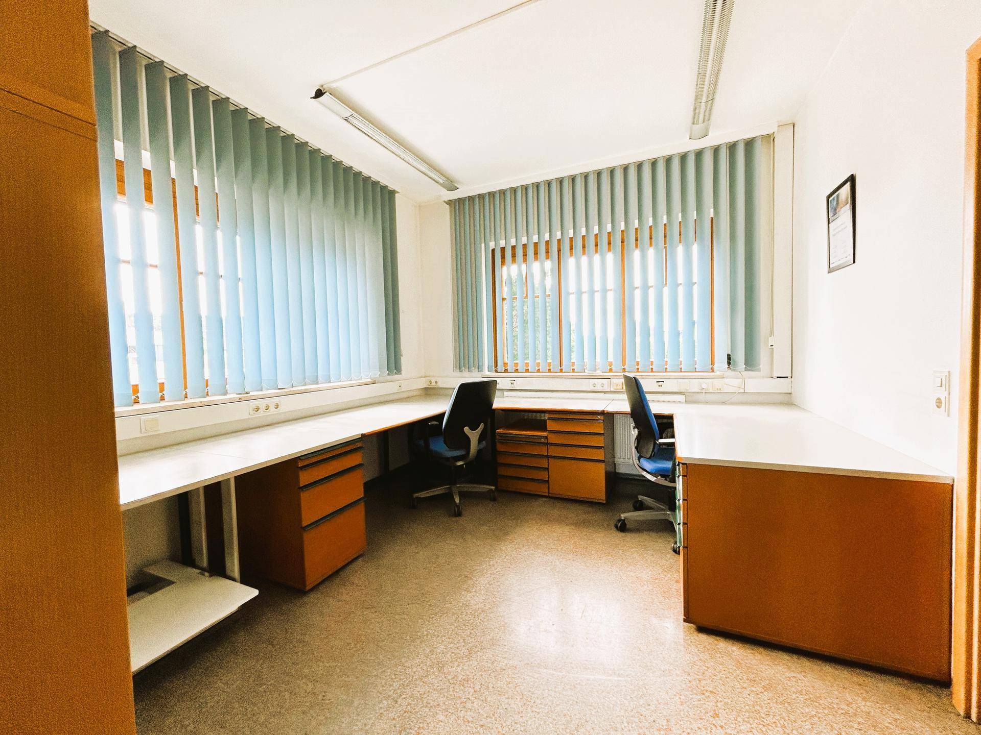 Helles Eckbüro mit großen Fenstern und Jalousien, ausgestattet mit zwei Arbeitsplätzen und viel natürlichem Licht.