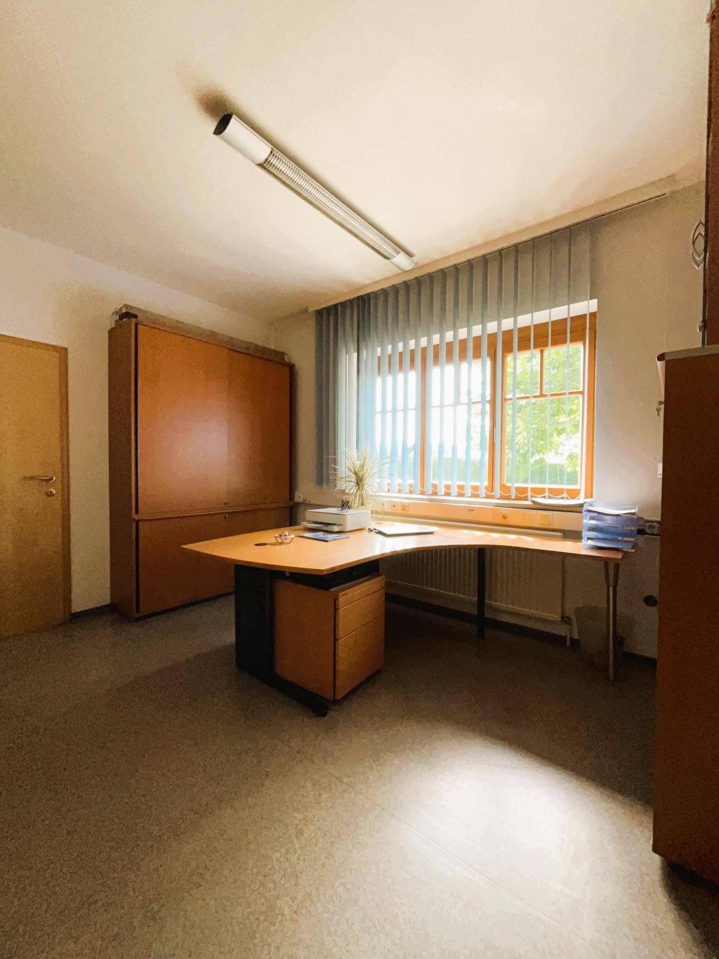Büro mit großem Schrank und Schreibtisch am Fenster, das viel Tageslicht hereinlässt.