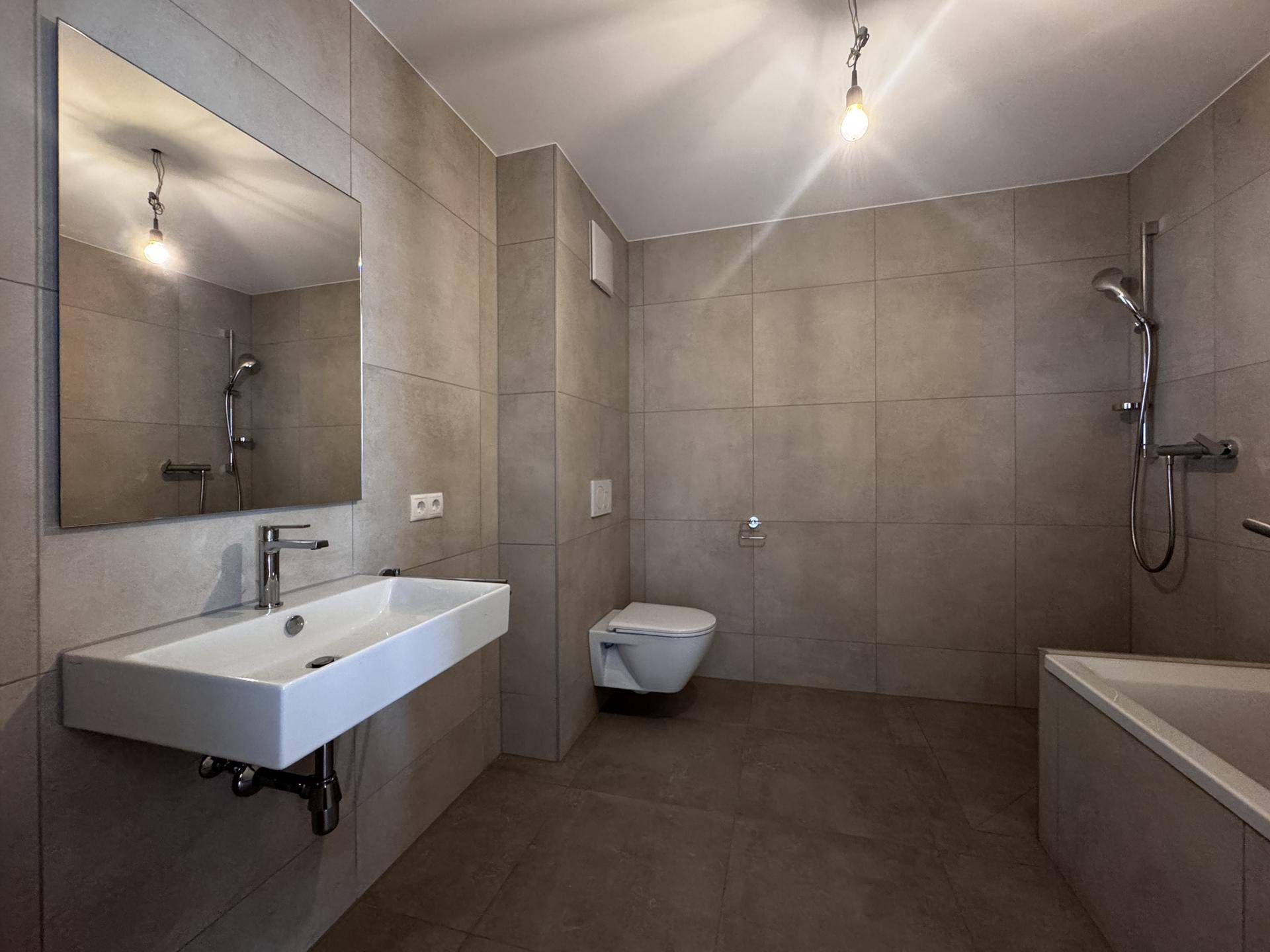 Modernes Badezimmer mit grauen Fliesen, wandhängendem WC, Waschtisch und Badewanne mit Duschfunktion.