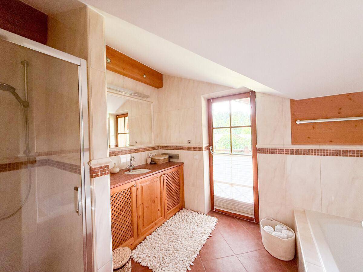 Badezimmer mit Duschkabine, Holzwaschtisch und Fenster, hell gefliest.