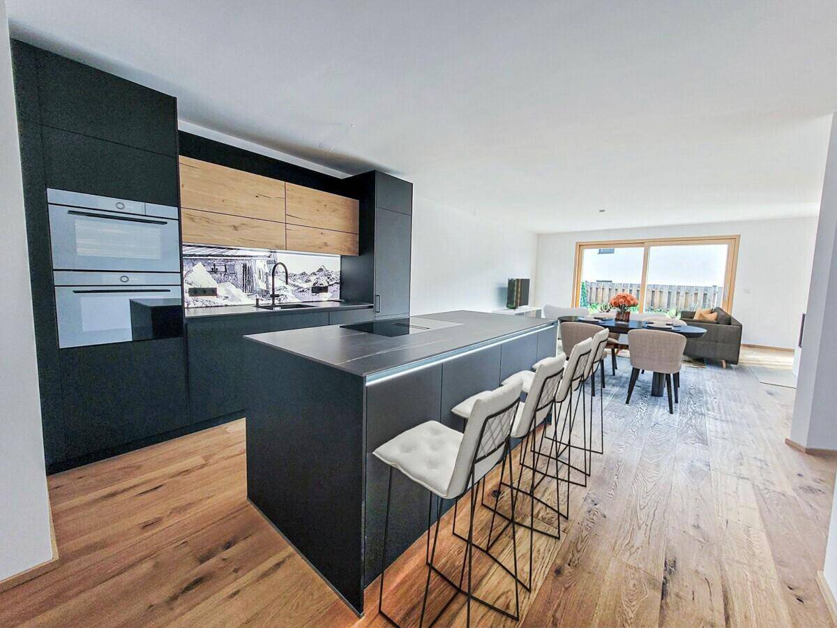 Moderne schwarze Küche mit Holzdetails, Kücheninsel und Essbereich im Hintergrund.