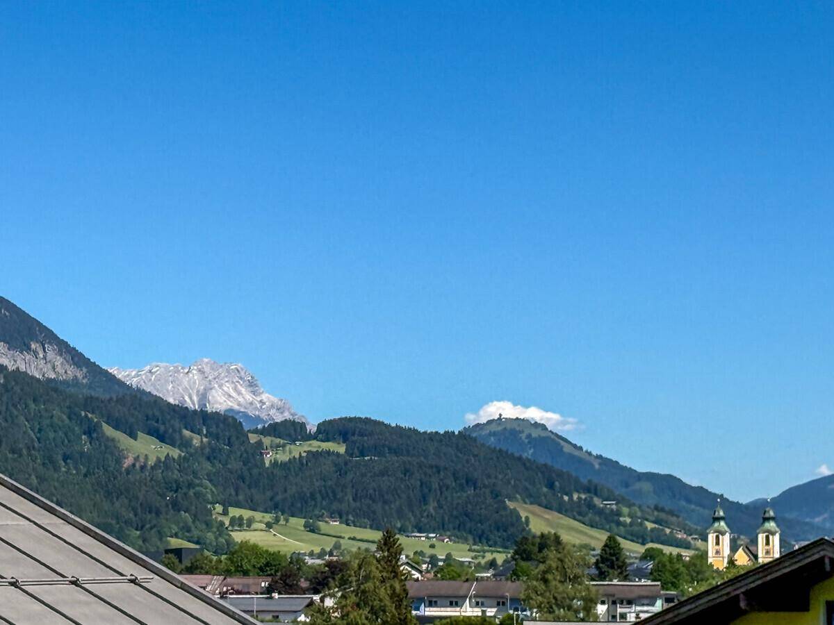 Weitläufiger Blick über eine Stadt mit Kirchtürmen und majestätischen Bergen im Hintergrund.