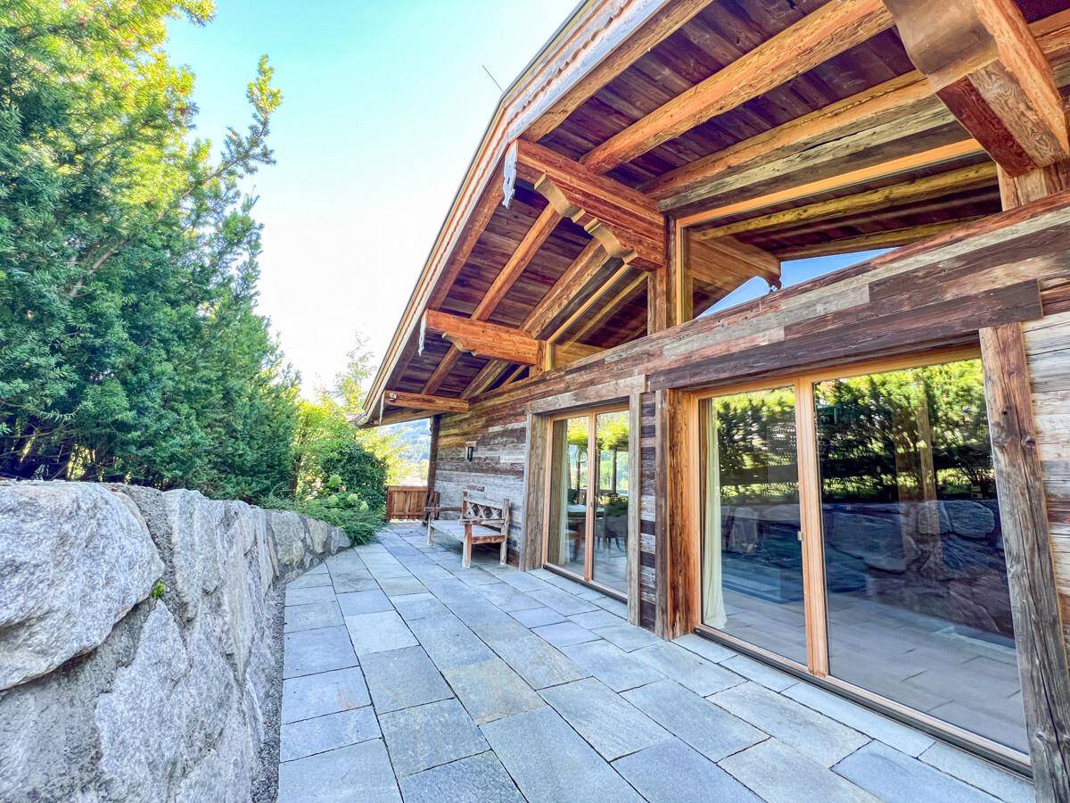 Große gepflasterte Terrasse mit Steinmauer und Zugang zum Holzchalet, umgeben von viel Grün.