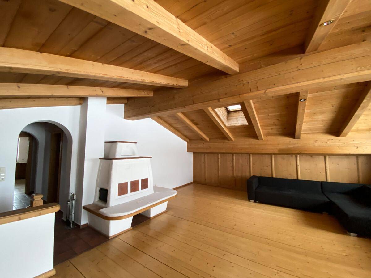 Gemütlicher Wohnbereich mit Holzbalken, Kachelofen und dunklem Sofa unter Dachschräge.