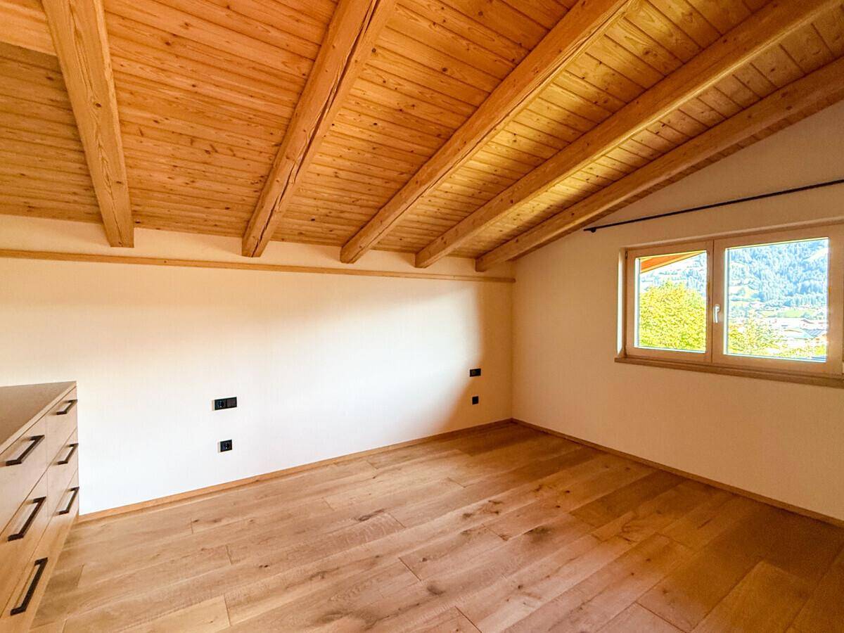 Helles Zimmer im Dachgeschoss mit Holzbalkendecke, Holzboden und Blick auf die grüne Umgebung.