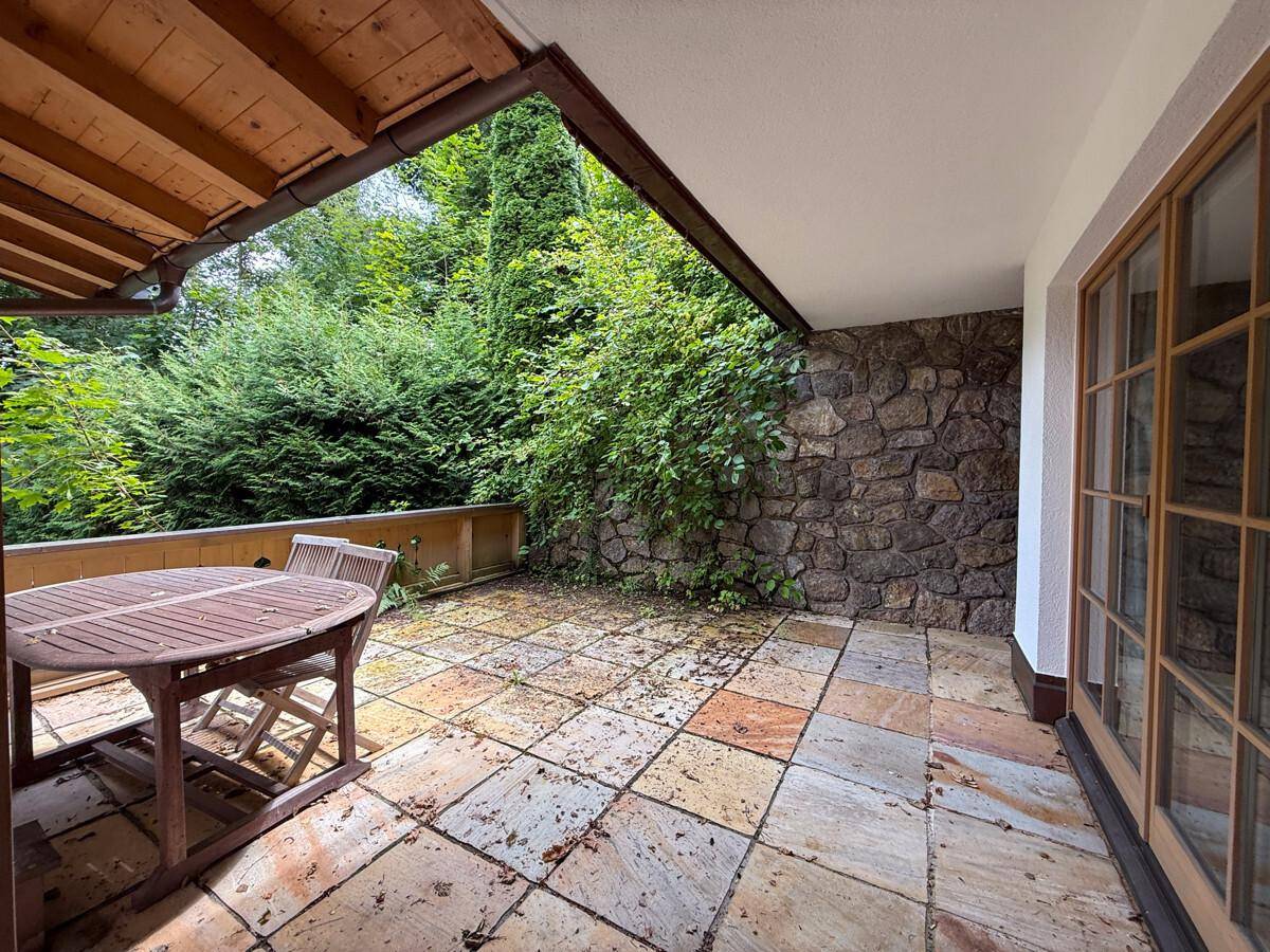 Geflieste Terrasse mit Holztisch, Stühlen und üppiger grüner Bepflanzung.