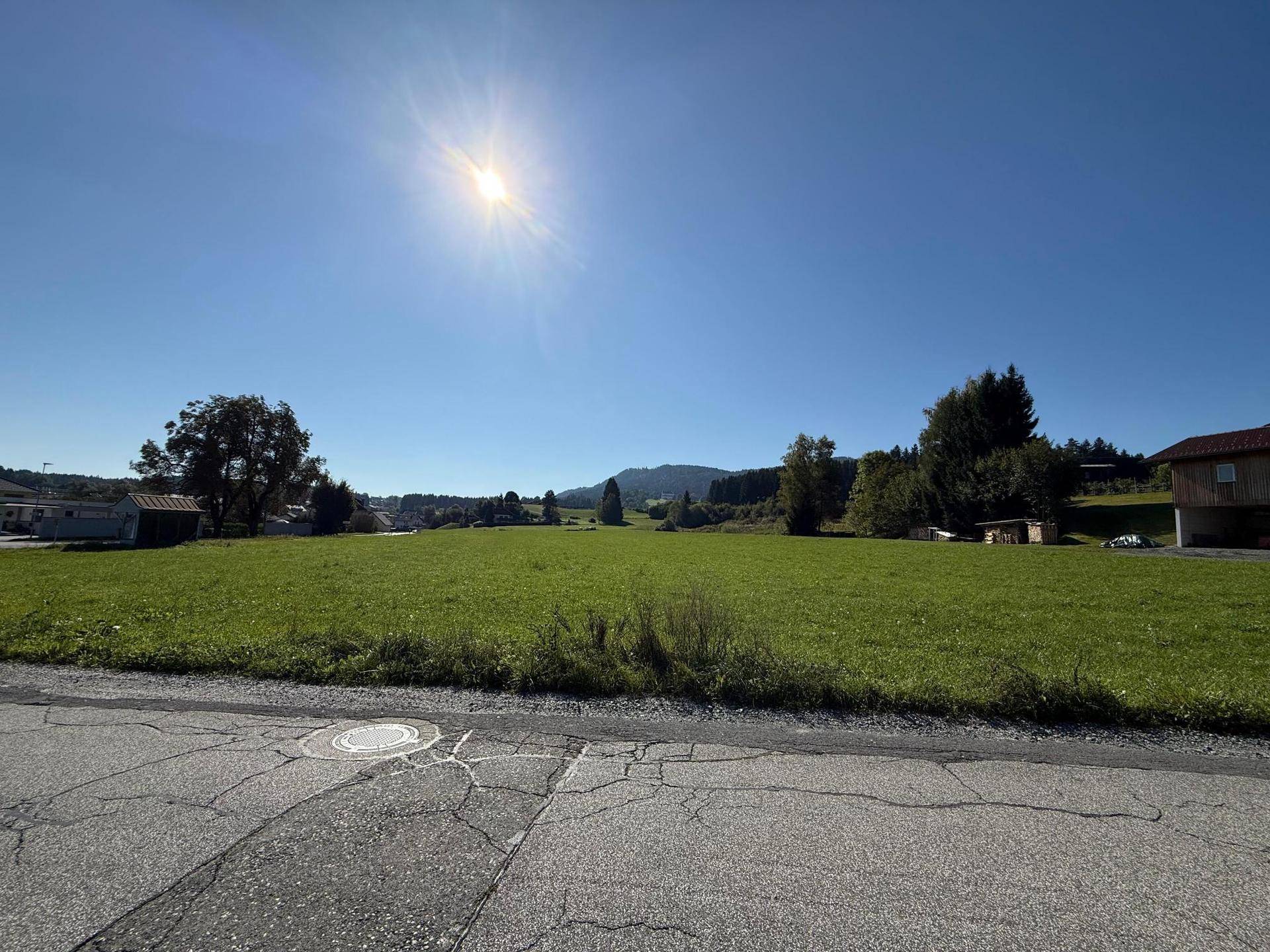 Offene Wiesenfläche unter strahlendem Sonnenschein mit Blick auf die ländliche Umgebung und ferne Hügel.