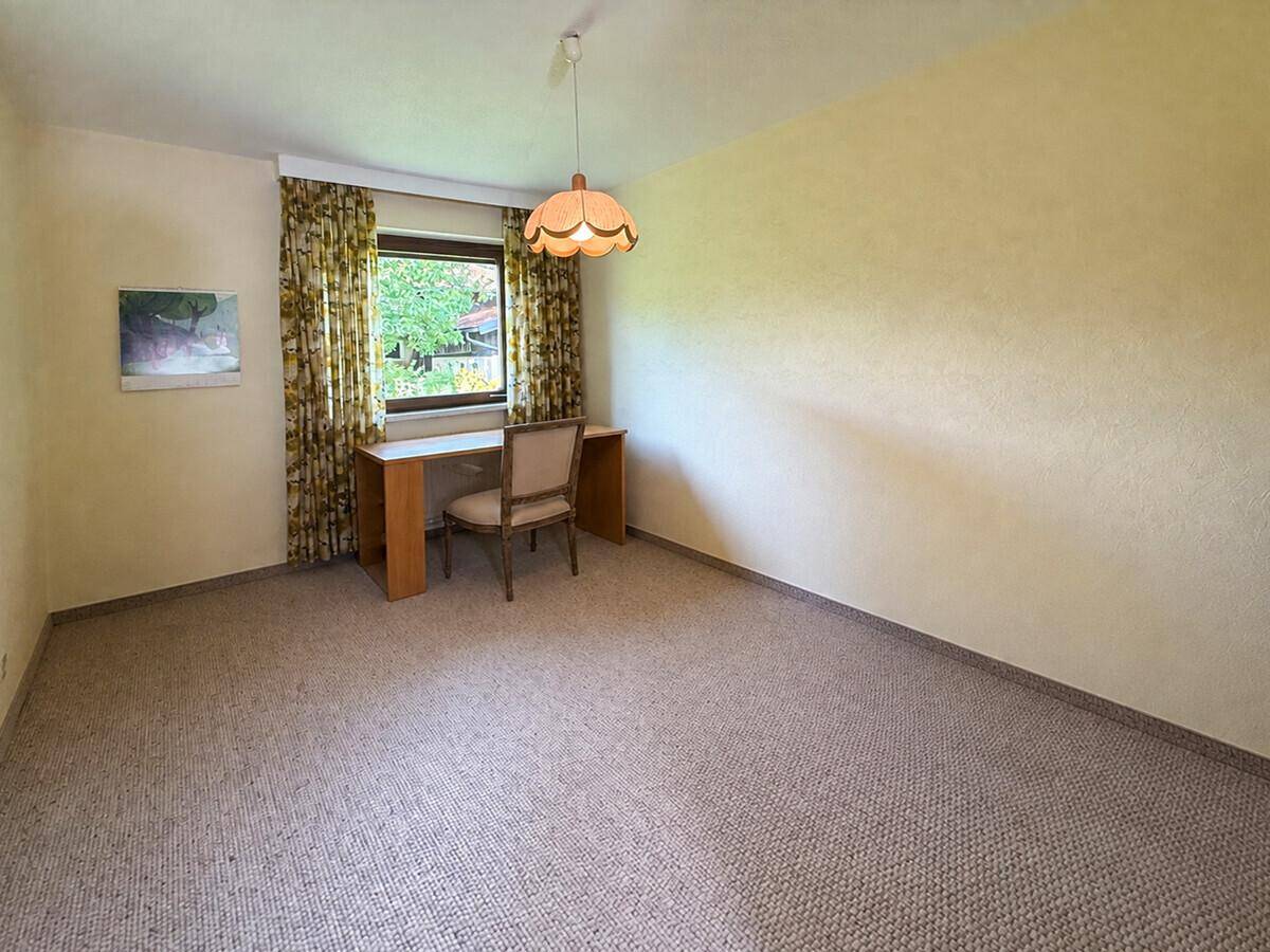 Helles Zimmer mit Teppichboden und einem Schreibtisch am Fenster.