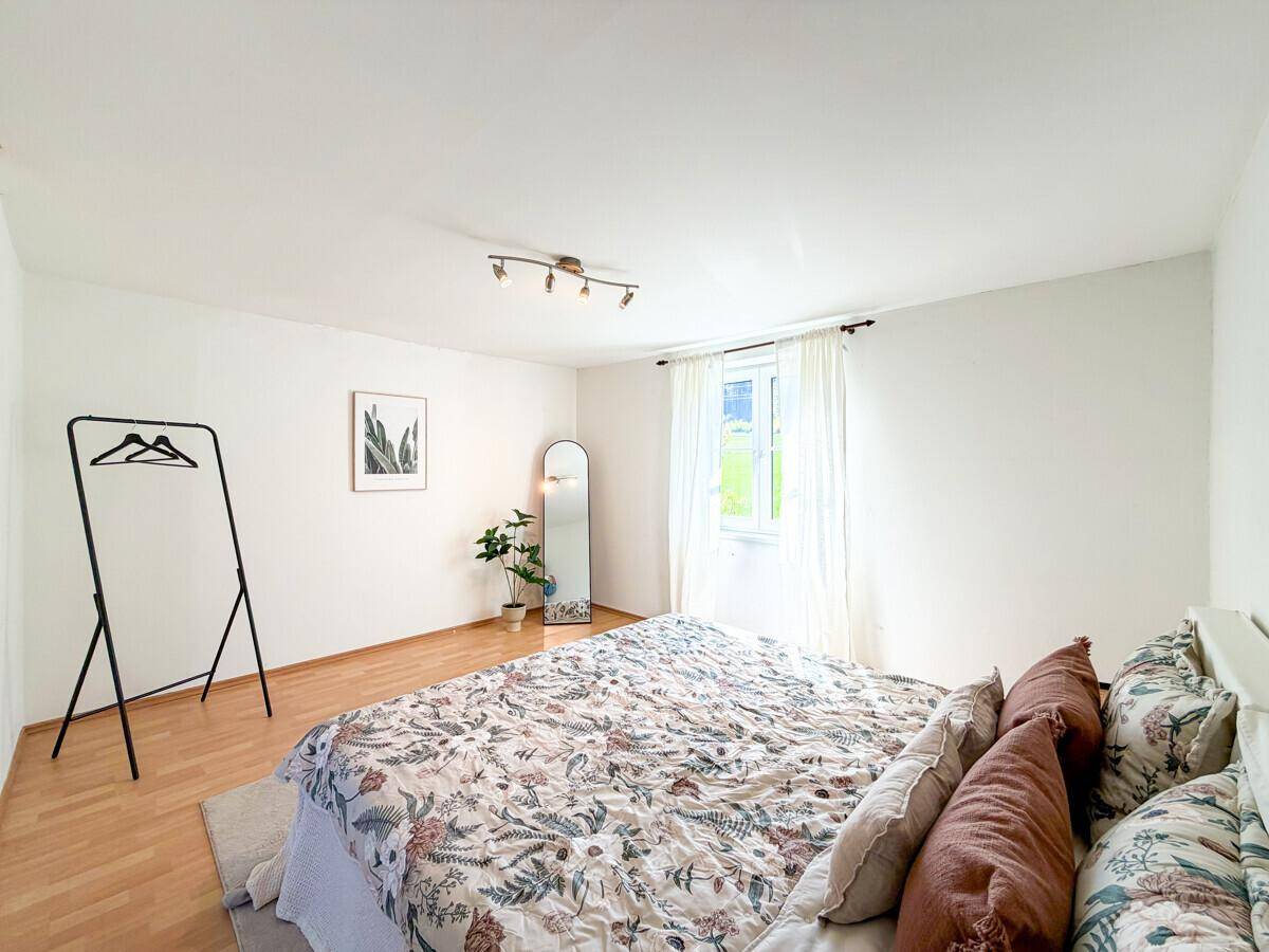 Schlafzimmer mit hellem Parkettboden, großem Bett und einem Fenster mit Blick ins Grüne.
