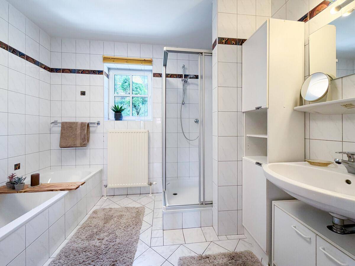 Badezimmer mit weißen Fliesen, Badewanne, Dusche und Fenster für Tageslicht.