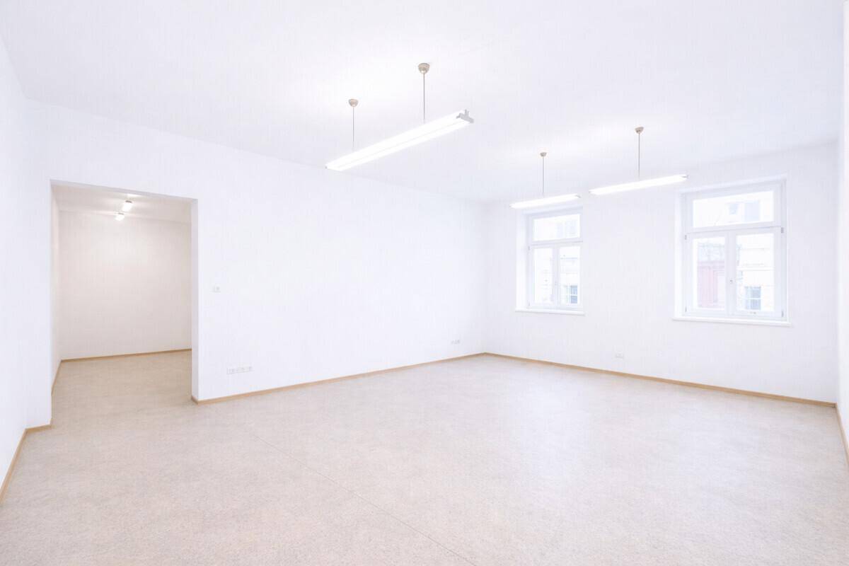 Großer, heller Raum mit weißen Wänden, Fliesenboden, zwei Fenstern und einem offenen Durchgang.