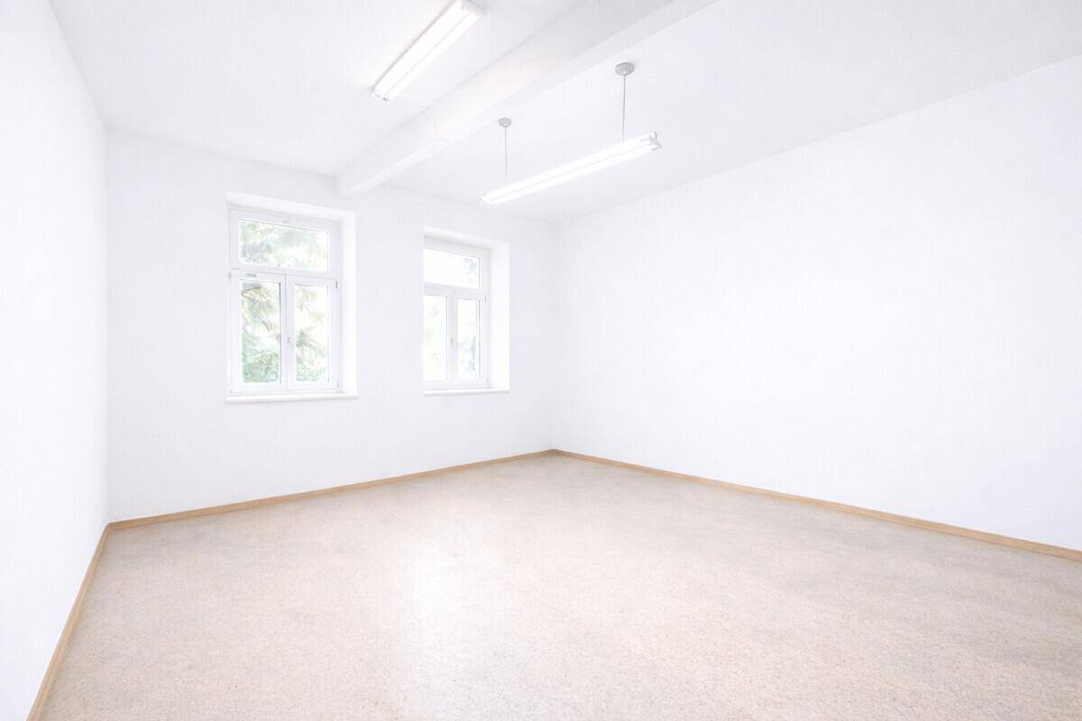 Heller, leerer Raum mit weißen Wänden, Fliesenboden und zwei Fenstern, die Tageslicht hereinlassen.