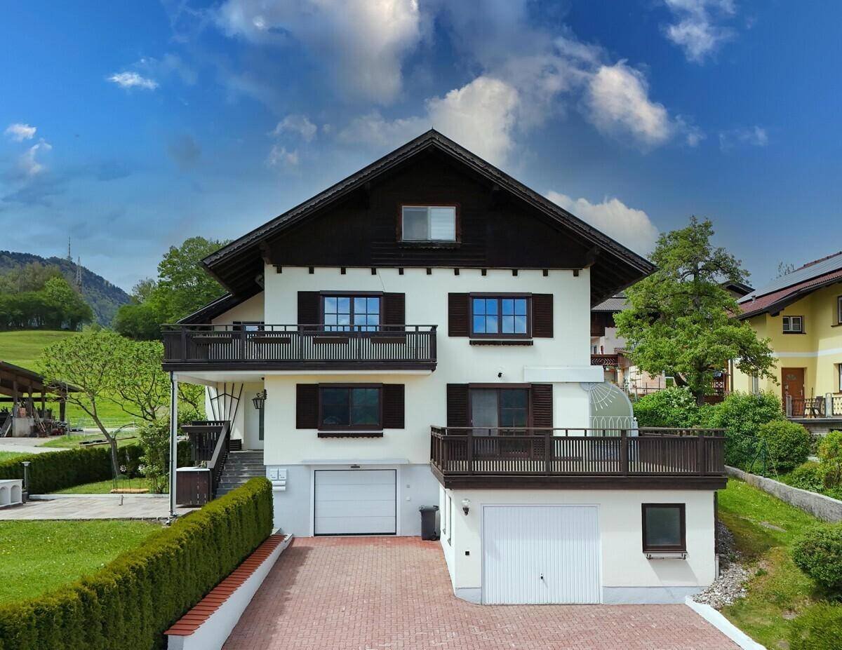 Frontansicht eines gepflegten Einfamilienhauses mit zwei Garagen, Balkonen und grüner Umgebung.