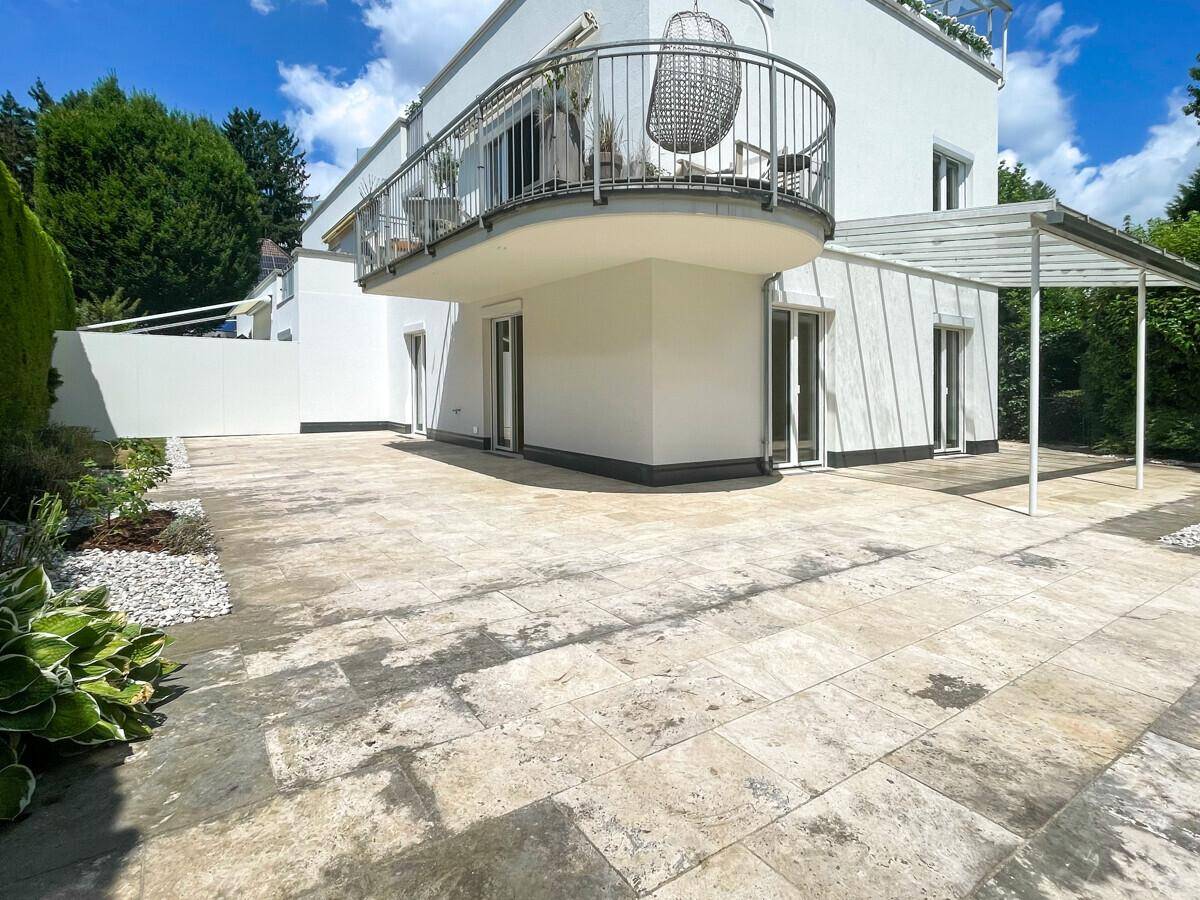 Große gepflasterte Terrasse mit Pergola und einem Balkon am modernen Gebäude.