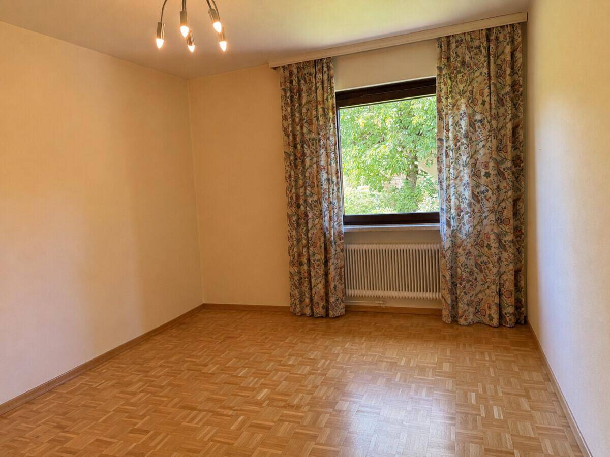 Leeres Zimmer mit Fischgrätparkett und großem Fenster mit Vorhängen.
