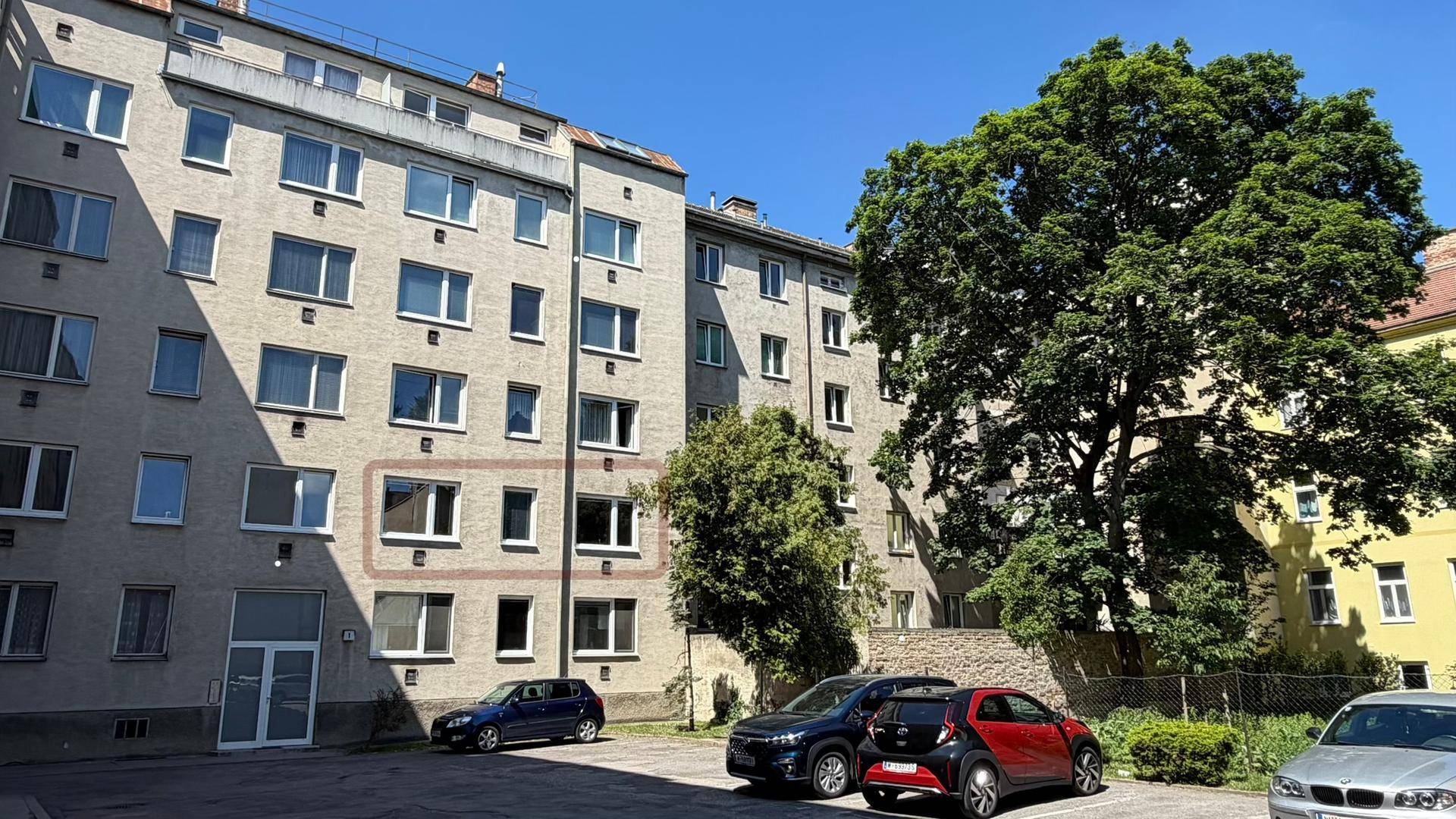 Fassade eines Mehrfamilienhauses mit geparkten Autos und großem Baum im Vordergrund, zeigt die Wohnumgebung.