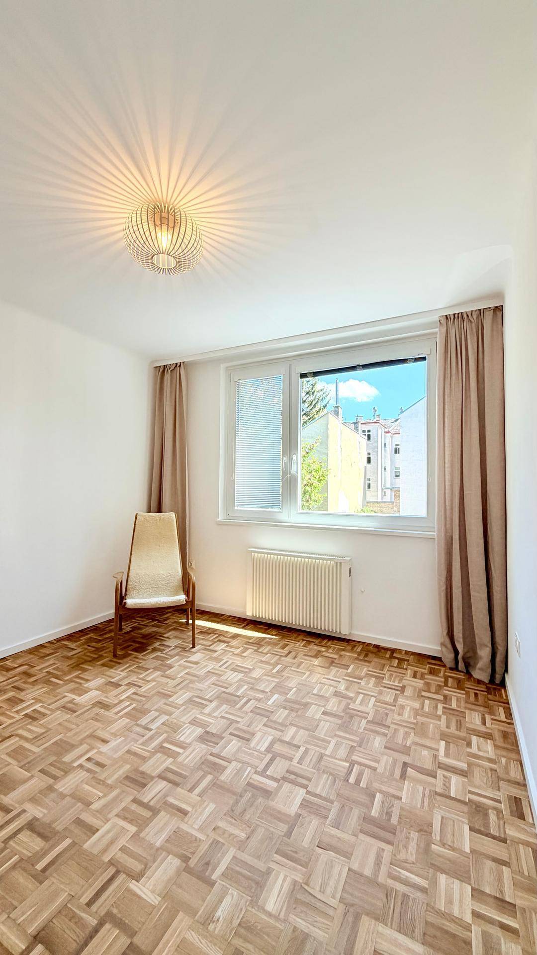 Helles Schlafzimmer mit Parkettboden, großem Fenster und einem einzelnen Sessel, bietet viel Tageslicht.