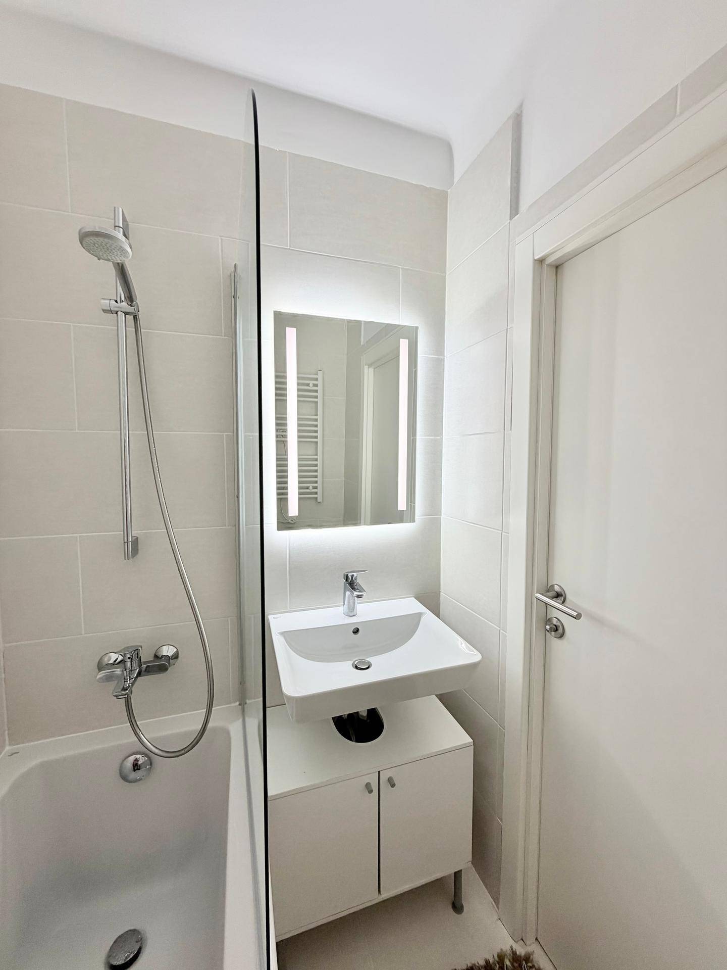 Modernes Badezimmer mit Badewanne, Duscharmatur, Waschbecken und beleuchtetem Spiegel, in hellen Tönen gehalten.
