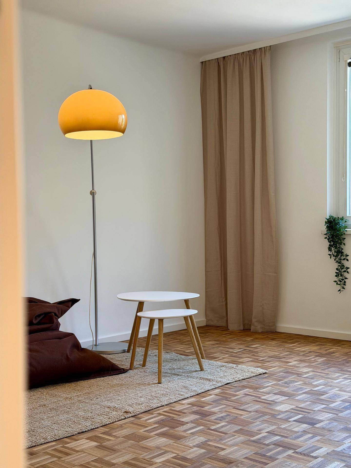 Detailansicht eines hellen Zimmers mit Stehlampe, Beistelltischen und Vorhängen am Fenster, gemütlich eingerichtet.