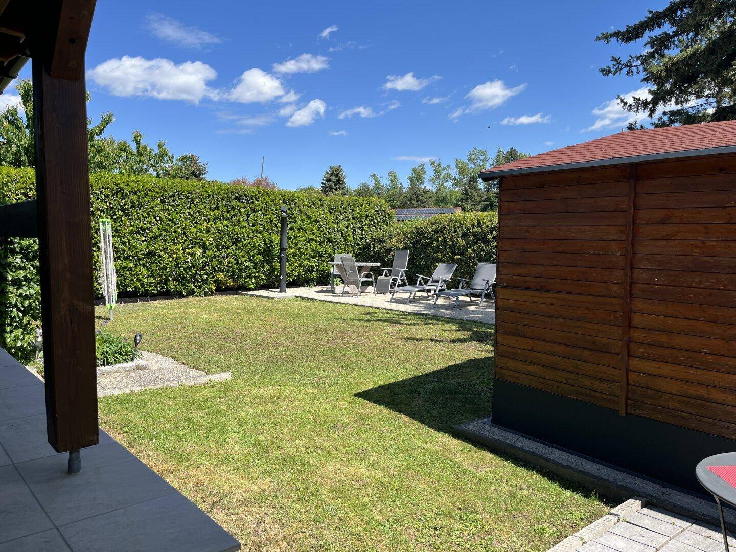 Gepflegter Gartenbereich mit Rasenfläche, Gartenhaus und Hecke unter blauem Himmel.
