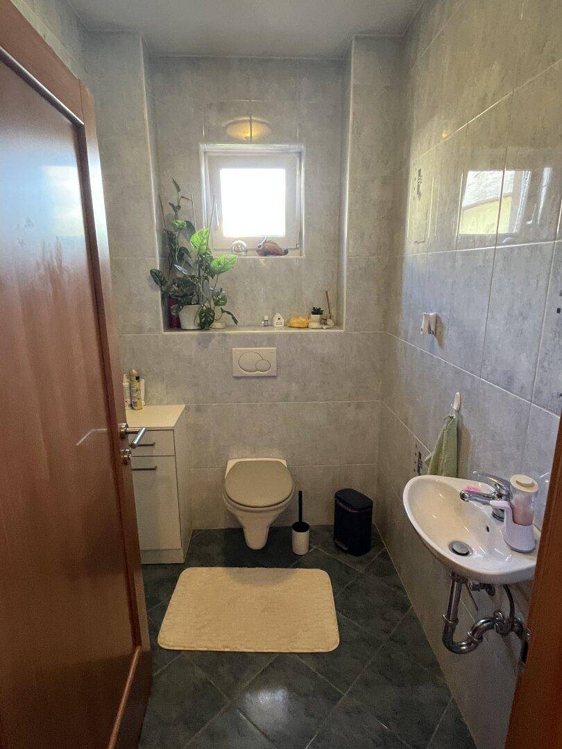 Kleines Gäste-WC mit Fenster, Wand-WC und Waschbecken in hellen Fliesen.