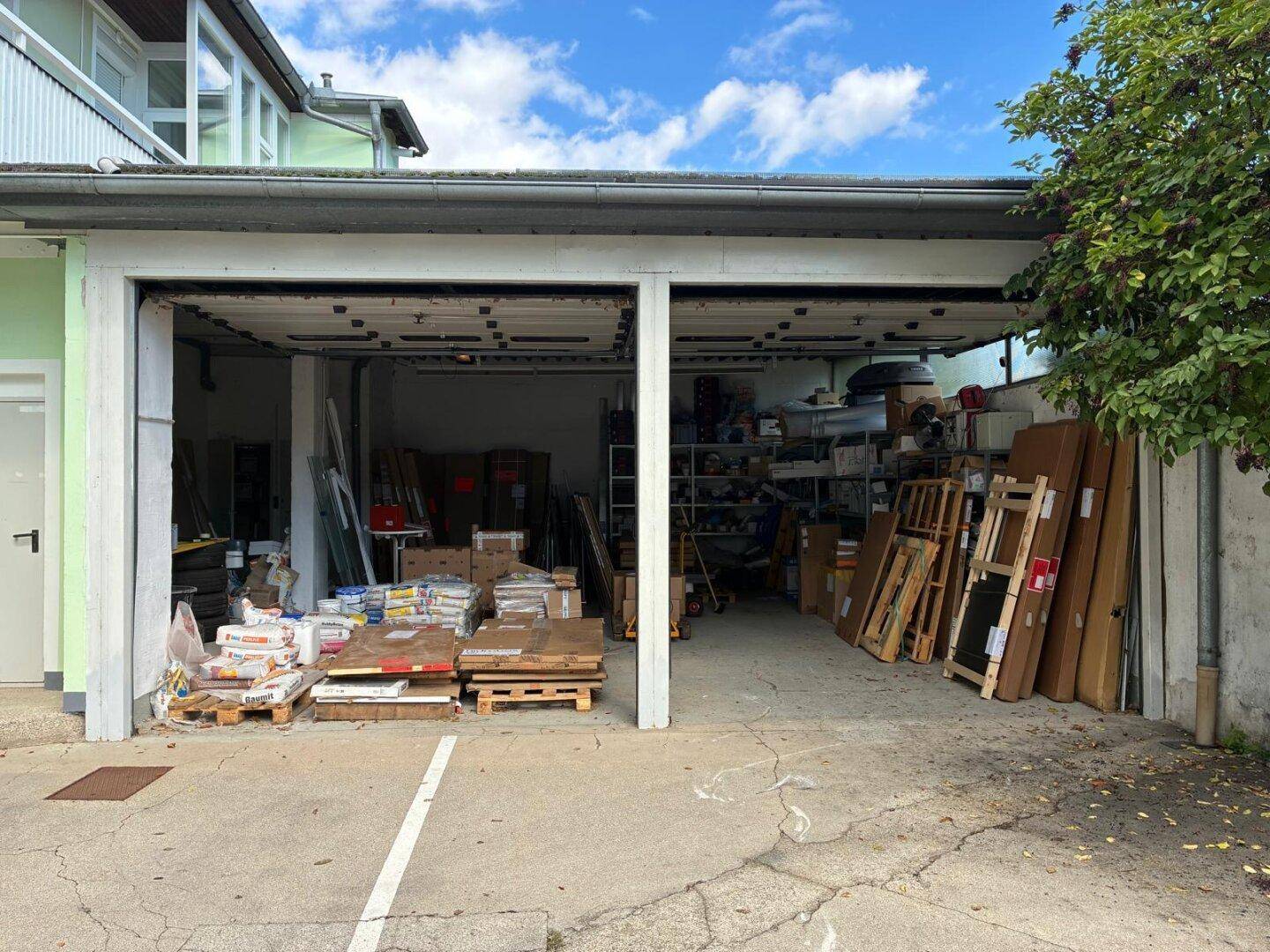 Zwei offene Garagentore zeigen unaufgeräumte Lagerbereiche, mit einem gepflasterten Vorplatz.