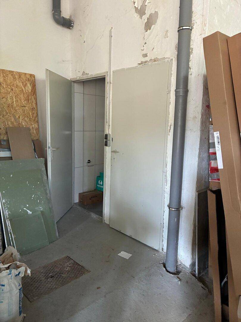 Innenansicht eines unaufgeräumten Raumes mit zwei offenen Türen, eine führt zu einem gefliesten Bereich.