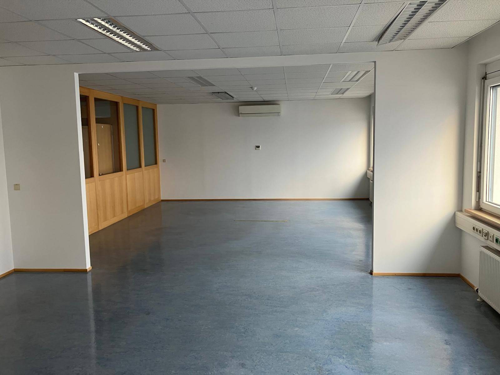 Helles Büro mit Klimaanlage, Holz-Glas-Trennwänden und grauem Linoleumboden.