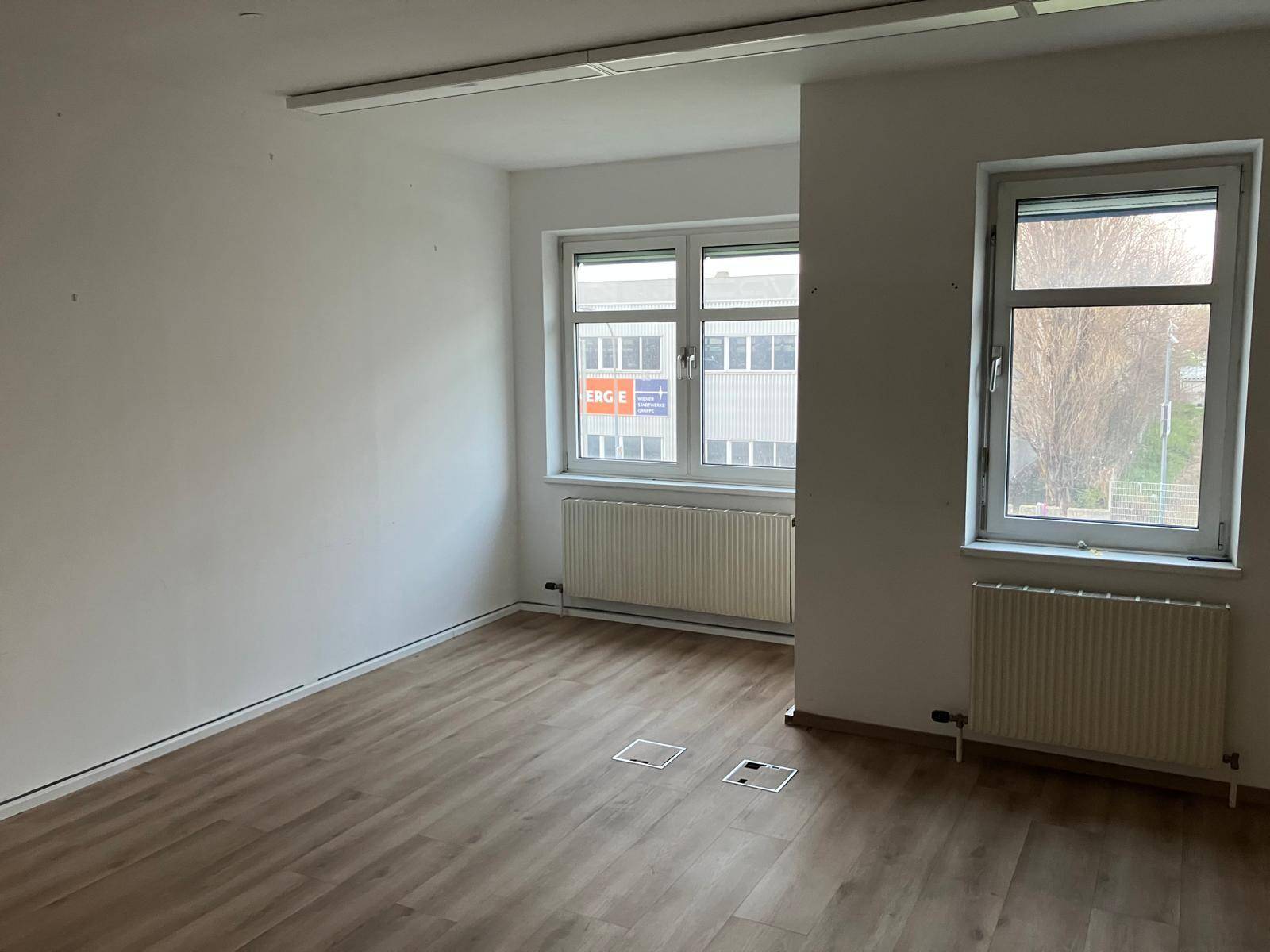 Leerer Büroraum mit zwei Fenstern, Laminatboden und Heizkörpern, bietet eine helle Arbeitsumgebung.