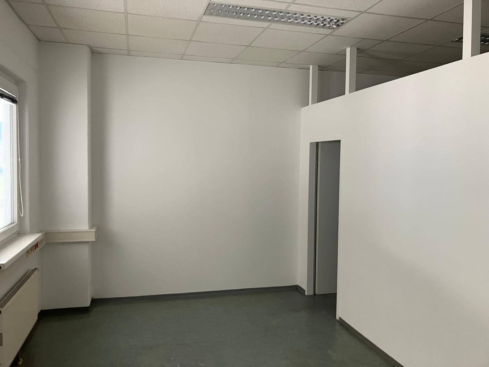 Büro mit weißen Wänden, einem Fenster und einem Durchgang zu einem weiteren Bereich, ausgestattet mit Vinylboden.