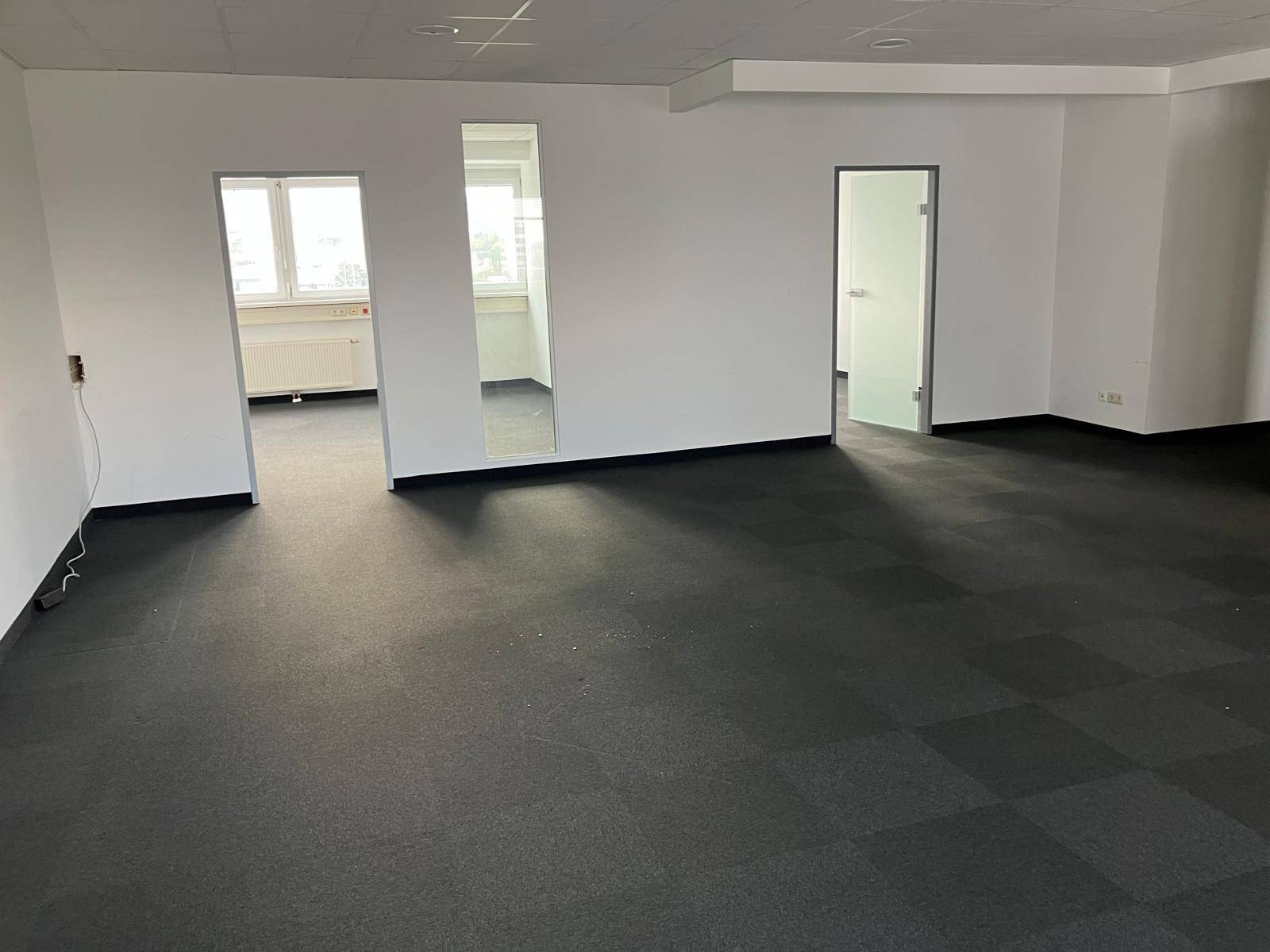 Helles, leeres Büro mit dunklem Teppichboden und Fenstern, die viel Tageslicht hereinlassen.