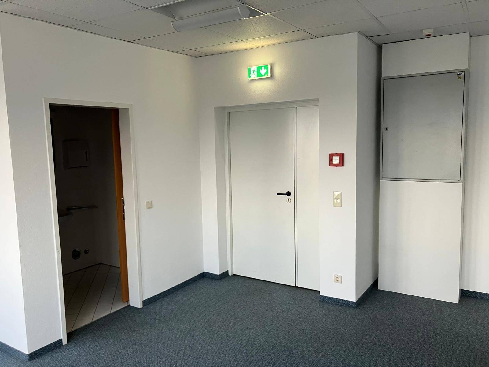Büroraum mit Teppichboden, weißen Wänden, Notausgang und Brandschutzvorrichtung.