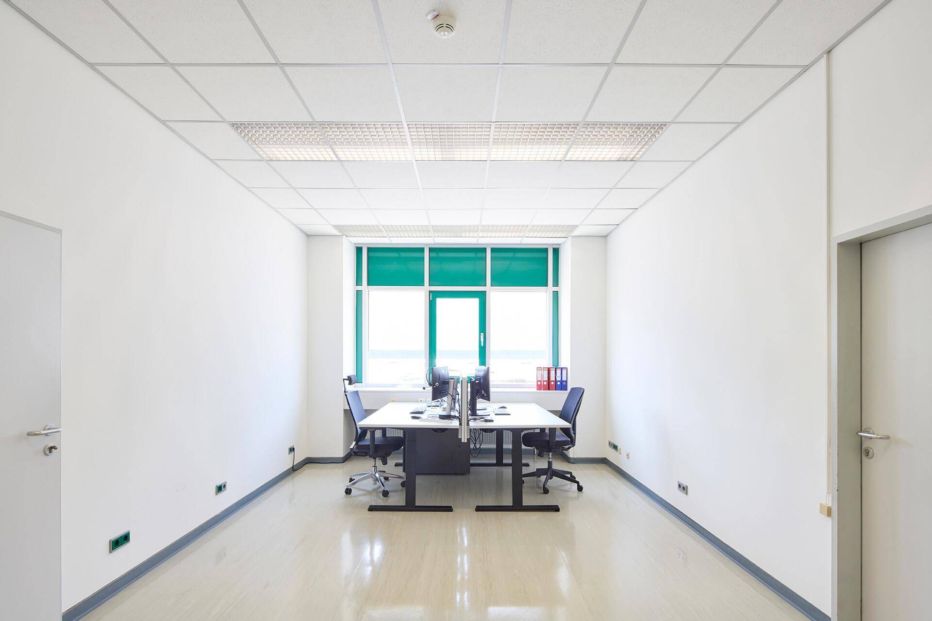 Helles, modernes Büro mit weißen Wänden, Rasterdecke und großen Fenstern mit grünen Jalousien, ausgestattet mit zwei Schreibtischen.