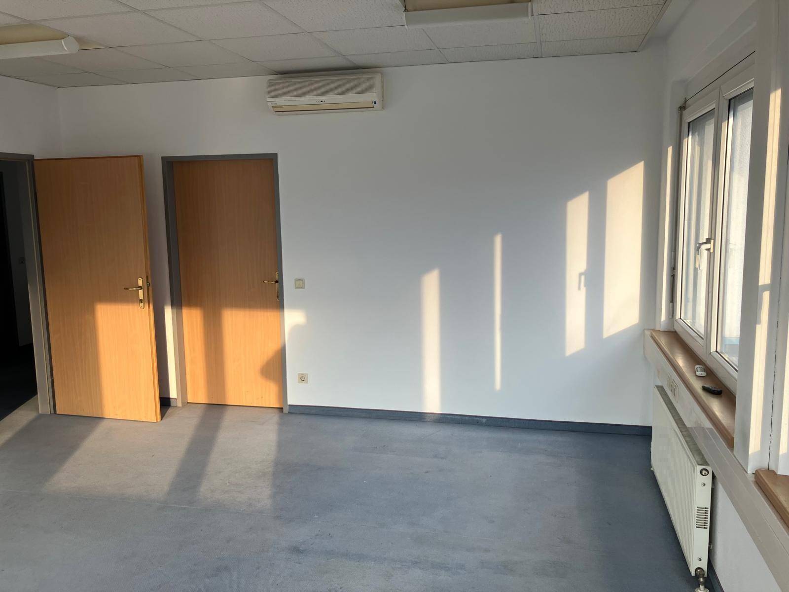 Geräumiger Büroraum mit Klimaanlage, großen Fenstern und hellem Vinylboden, beleuchtet von natürlichem Sonnenlicht.