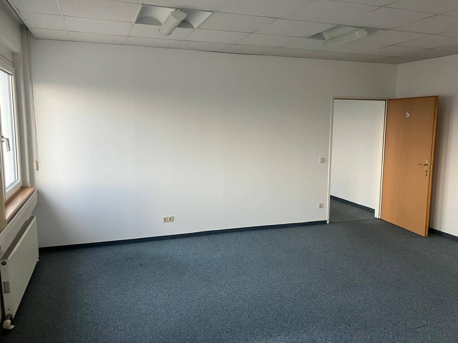 Leerer Büroraum mit Teppichboden, weißen Wänden, einer Tür und einem Fenster.
